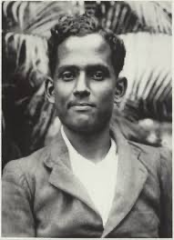 An outdoor photograph of Jatindra Nath Das as a young man in a light jacket and collarless shirt, standing in front of foliage.