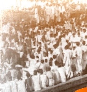 An aged photograph of a very large crowd of people, many in white clothing, gathered densely along a raised structure.